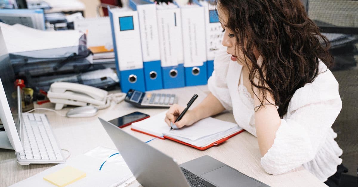 An office worker takes notes while working at a desk with a laptop and documents.