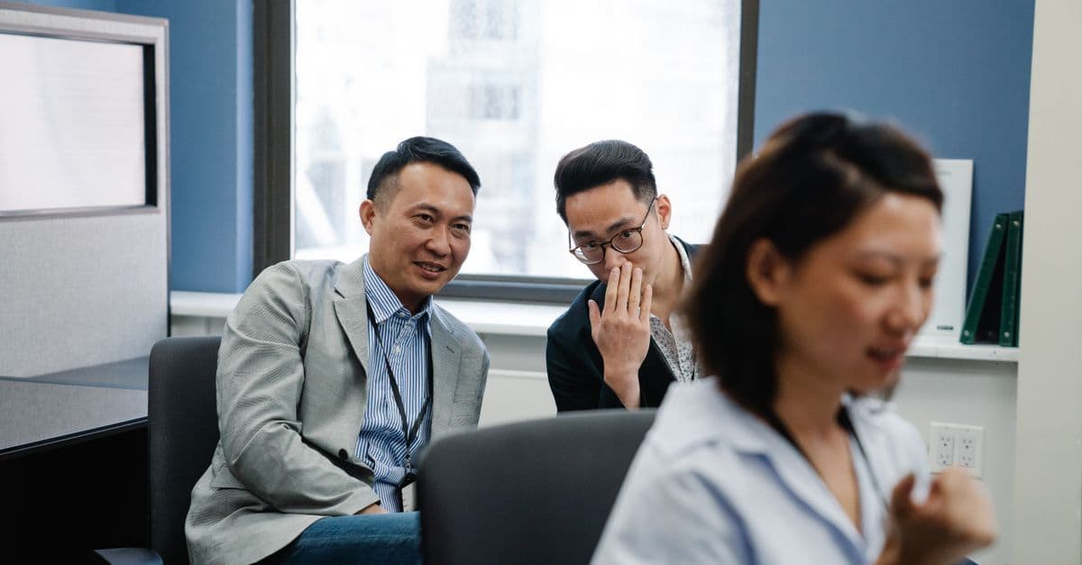 Two male colleagues sharing a secret in an office while a female coworker works nearby.