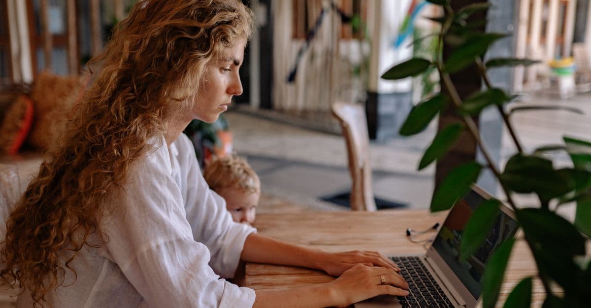 Woman with curly hair working on a laptop at home, with a child nearby, showcasing remote work lifestyle.