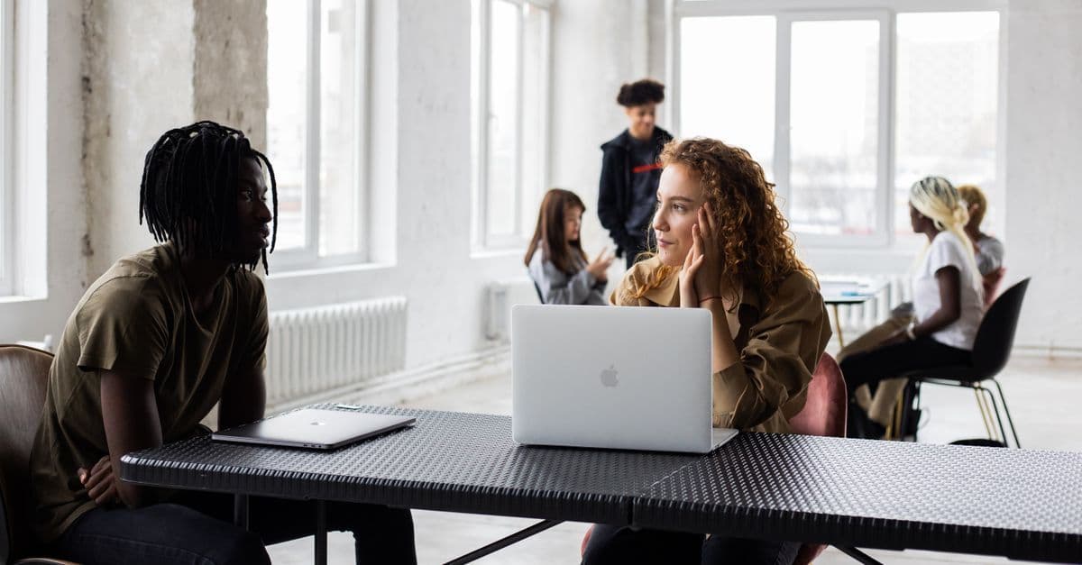Concentrated young multiethnic coworkers in casual clothes sitting at table with laptops and discussing project details in light loft workspace