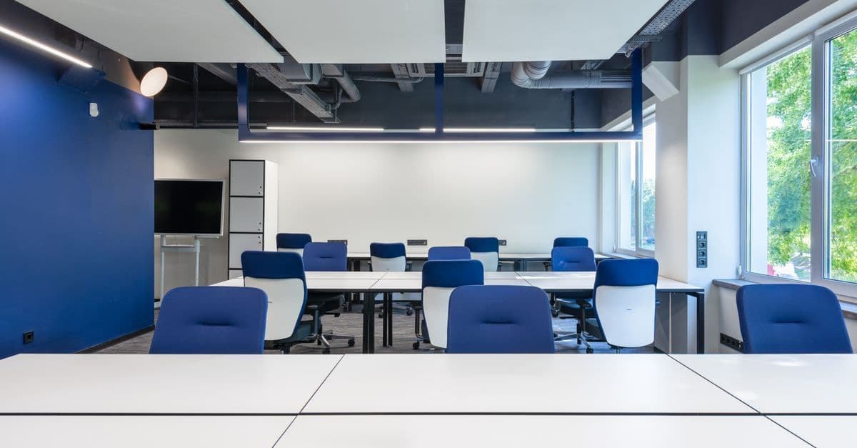Various white desks with blue chairs placed in modern spacious workspace with big window and monitor in contemporary business center