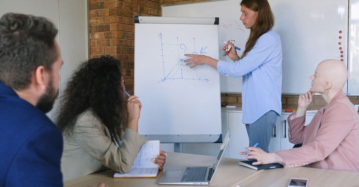 Group of pensive coworkers sitting at wooden table with gadgets and documents and listening presentations of colleague drawing graph in flipchart in modern office workspace in daytime