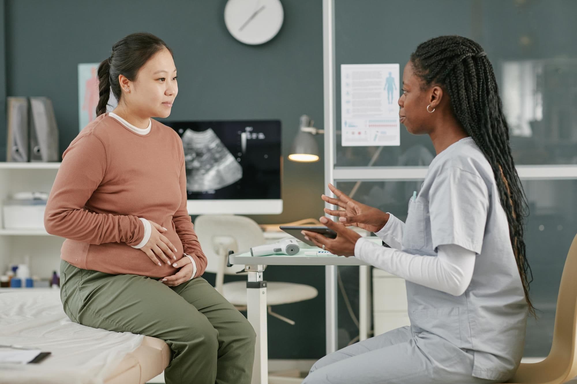 Pregnant young adult asian woman consulting black female doctor in clinic