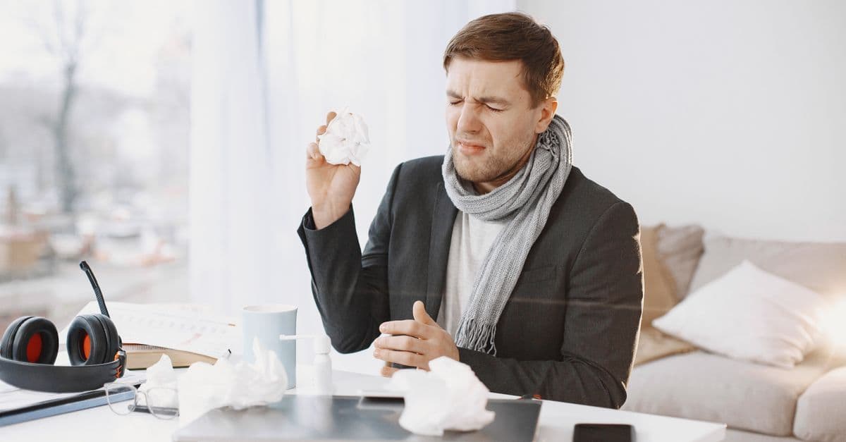 A sick man wearing a scarf sitting at his desk, holding a tissue, displaying signs of discomfort.