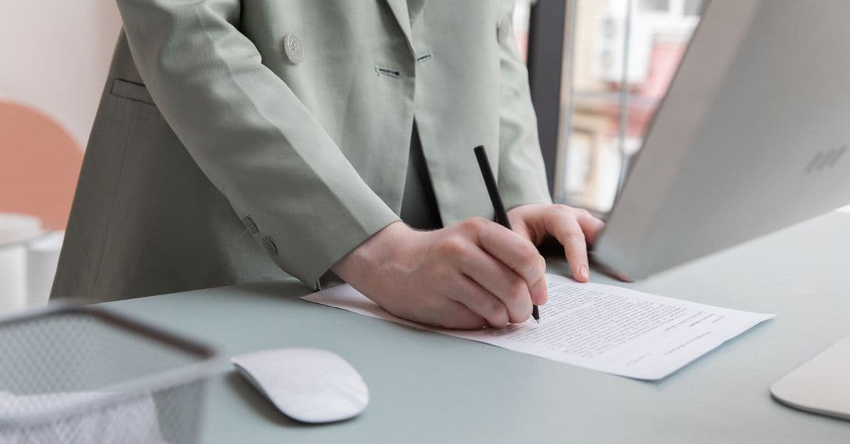 Unrecognizable worker standing at table with computer while taking notes in document while working in office