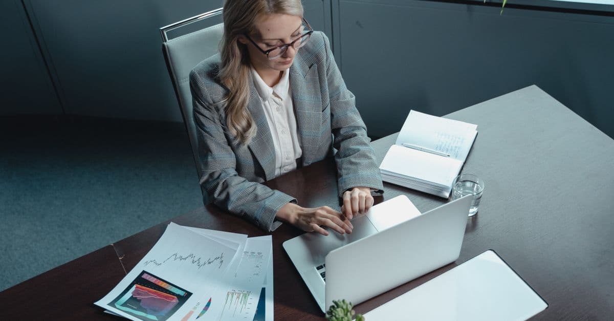 Blonde woman in a blazer working on a laptop in a modern office with documents.