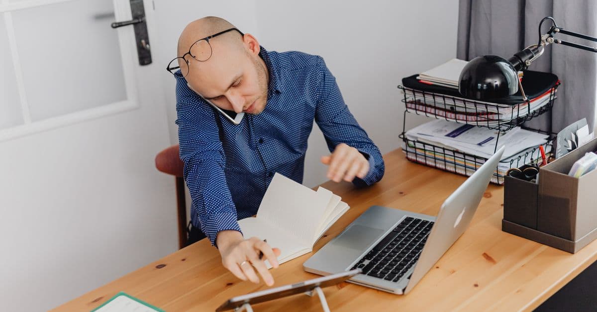 A man multitasking with phone, laptop, and notebook on his desk at home office.