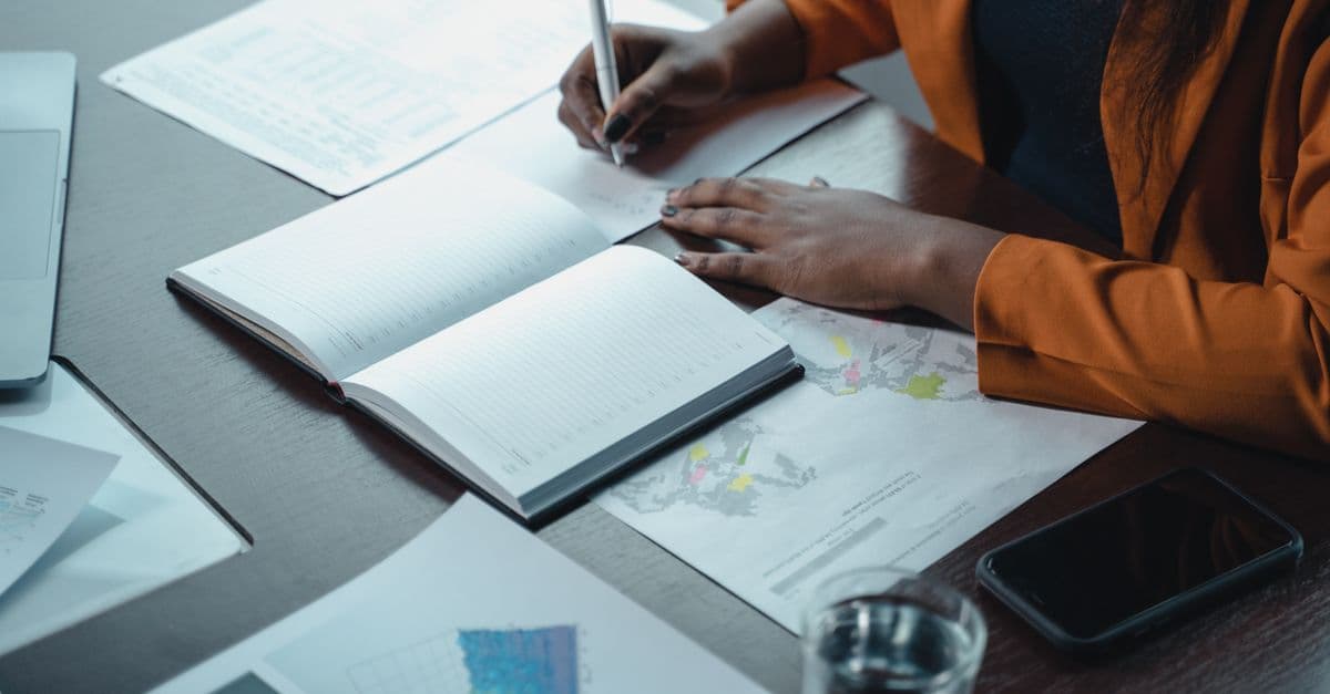 A businesswoman writes in a notebook while reviewing charts and graphs in an office setting.
