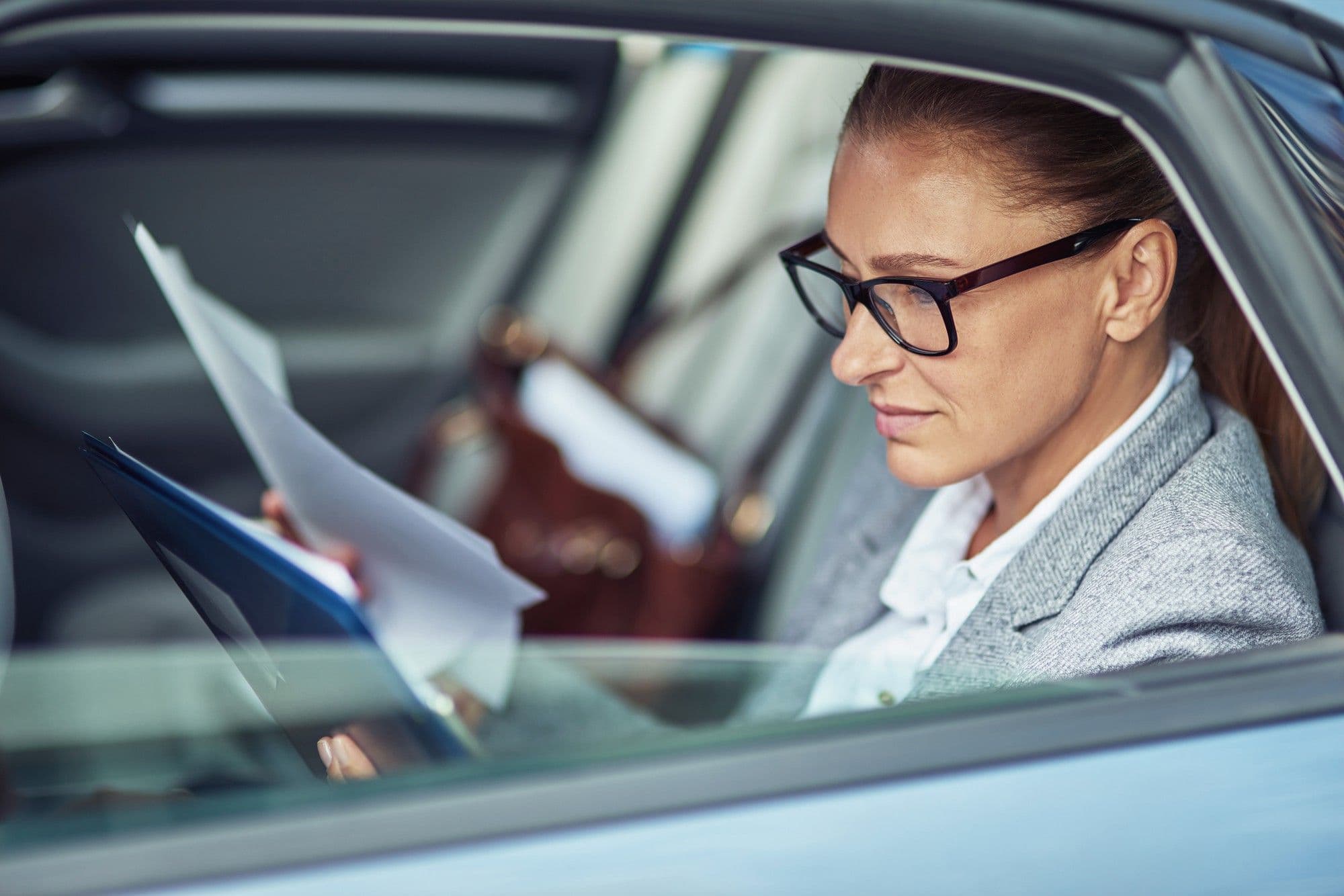 Side view of a focused middle aged business woman wearing eyeglasses analyzing documents while sitting on back seat in the car. Transportation and vehicle concept, business people