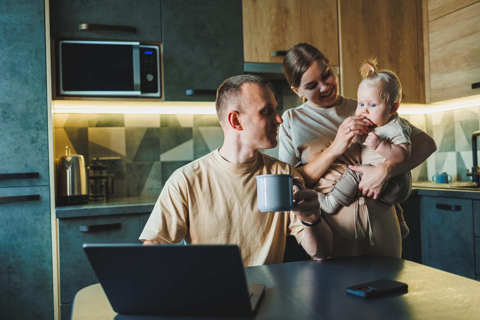 A young couple with a child in the kitchen A man works at home on a laptop and his wife with a child in her arms is standing next to him Family relationships